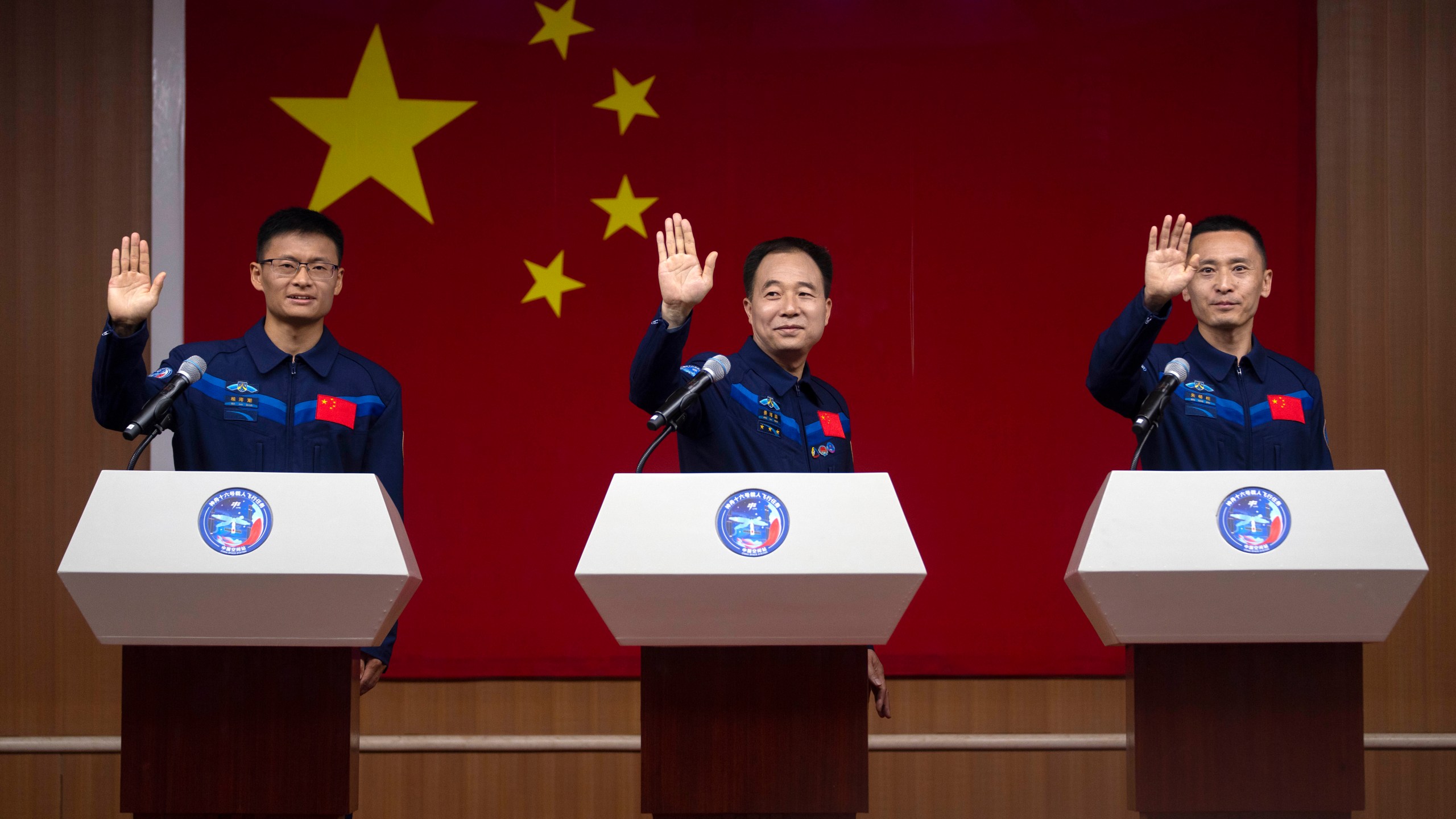 Chinese astronauts for the upcoming Shenzhou-16 mission, from left, Gui Haichao, Jing Haipeng, and Zhu Yangzhu wave as they stand behind glass during a meeting with the press at the Jiuquan Satellite Launch Center in northwest China on Monday, May 29, 2023. China's space program plans to land astronauts on the moon before 2030, a top official with the country's space program said Monday. (AP Photo/Mark Schiefelbein)