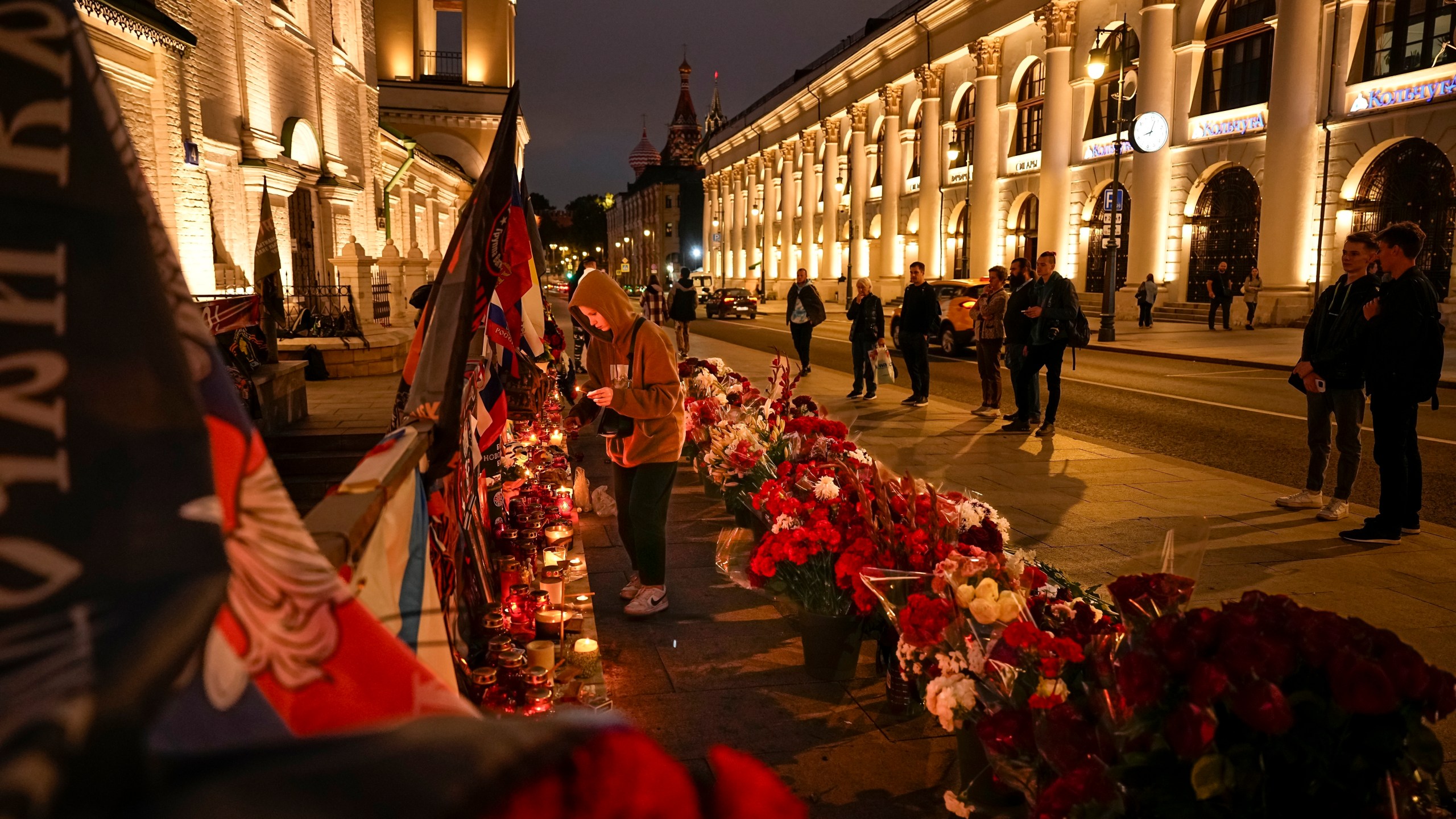 A young woman lights a candle as others stand at an informal street memorial with flowers and lit candles for Wagner Group's military group members killed in a plane crash, near the Kremlin, in the background, in Moscow, Russia, Tuesday, Aug. 29, 2023. The Kremlin says Russian President Vladimir Putin isn't planning to attend the funeral of Yevgeny Prigozhin, who died last week in a plane crash two months after launching his brief rebellion. (AP Photo/Alexander Zemlianichenko)