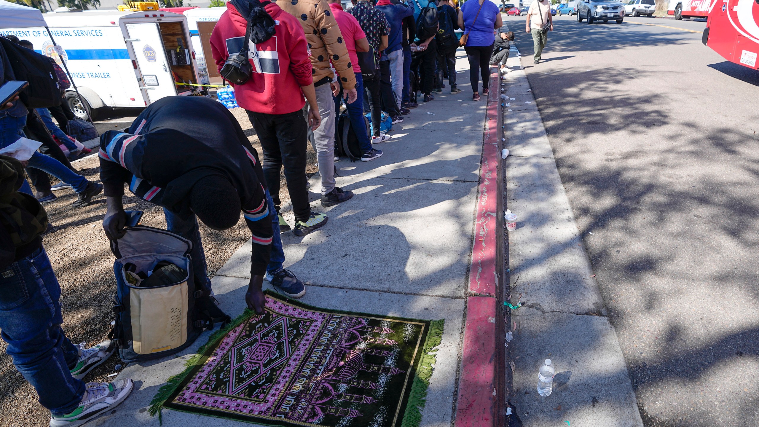 A migrant picks up his rug after prayer as he waits for a bus to the airport Friday, Oct. 6, 2023, in San Diego. San Diego's well-oiled system of migrant shelters is being tested like never before as U.S. Customs and Border Protection releases migrants to the streets of California's second-largest city because shelters are full. (AP Photo/Gregory Bull)