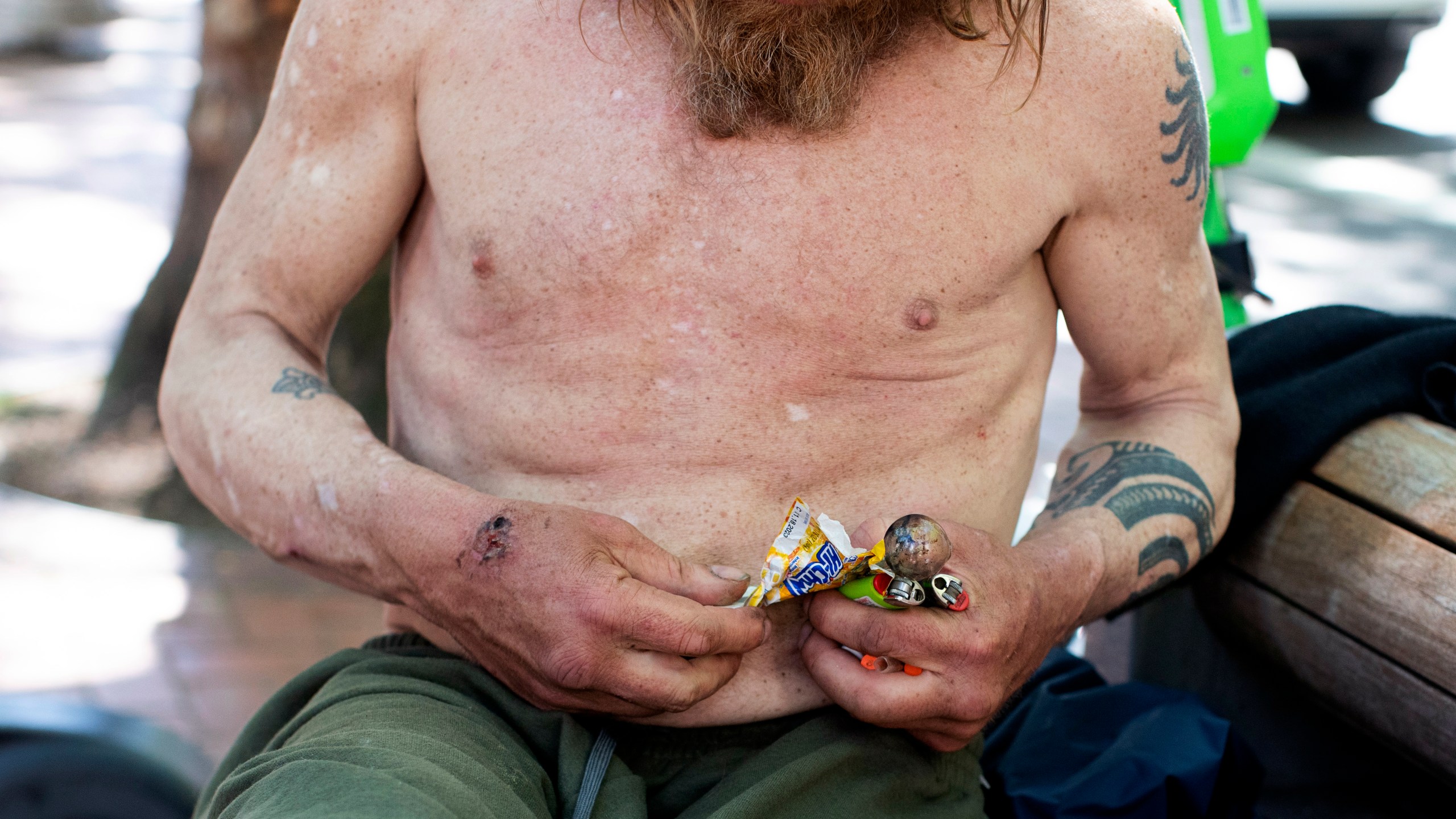 A man holding a glass pipe and two lighters struggles to wrestle a piece of candy from a wrapper while sitting on a bench in downtown Portland, Ore. on Thursday, May 18, 2023. Three years ago, nearly two-thirds of Oregon voters approved a ballot measure decriminalizing illicit drugs, backing the idea that addiction treatment is more effective than jail. But now, public drug use in cities such as Portland and a surge in fentanyl overdose deaths have created a backlash against the first-in-the-nation law. (Beth Nakamura/The Oregonian via AP)