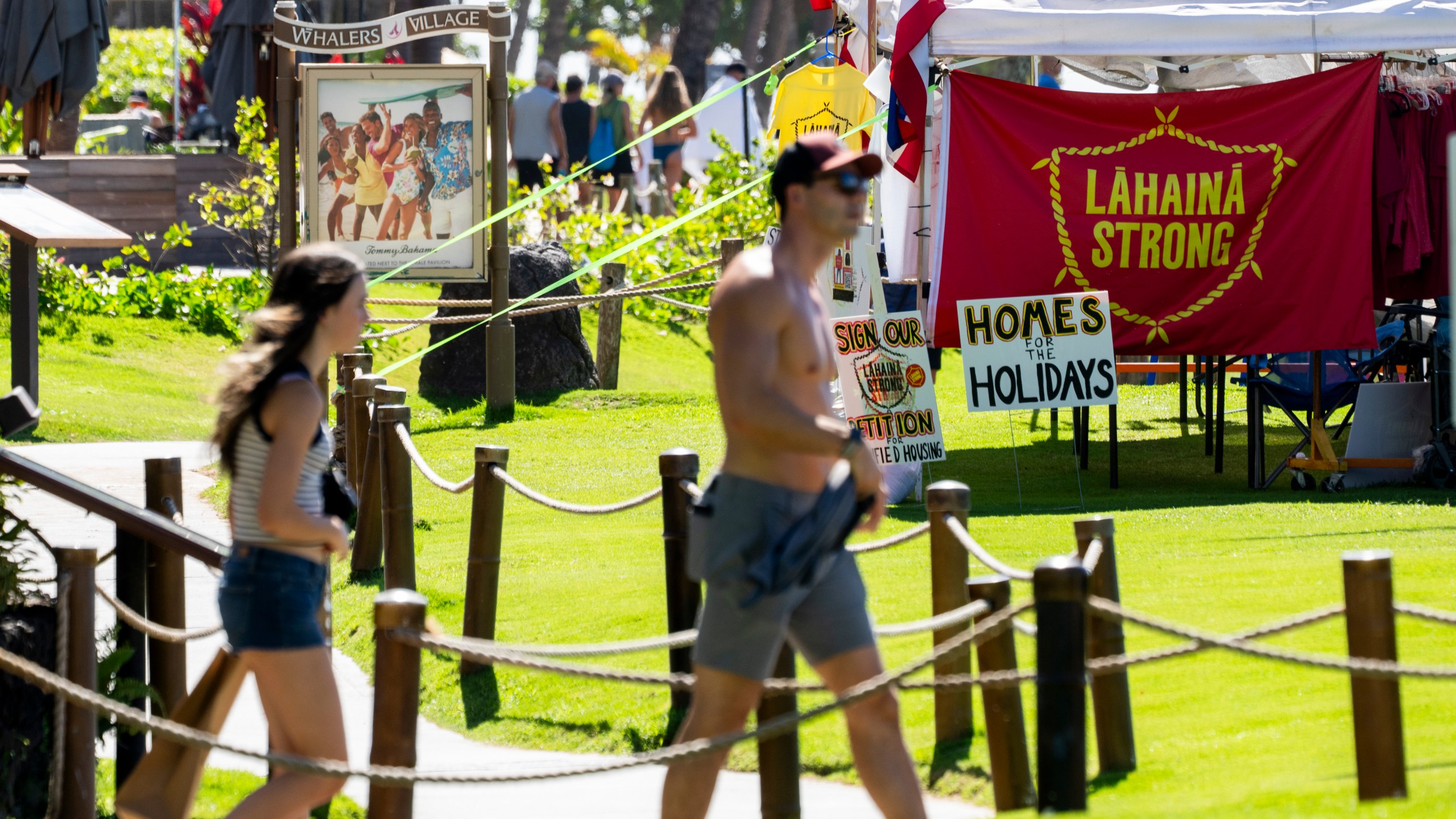 People walk by a tent with a "Lahaina strong" flag, Wednesday, Dec. 6, 2023, at Whalers Village on Kaanapali Beach in Lahaina, Hawaii. Residents and survivors still dealing with the aftermath of the August wildfires in Lahaina have mixed feelings as tourists begin to return to the west side of Maui, staying in hotels still housing some displaced residents. A group of survivors is camping on the resort beach to protest and raise awareness for better long-term housing options for those displaced. (AP Photo/Lindsey Wasson)