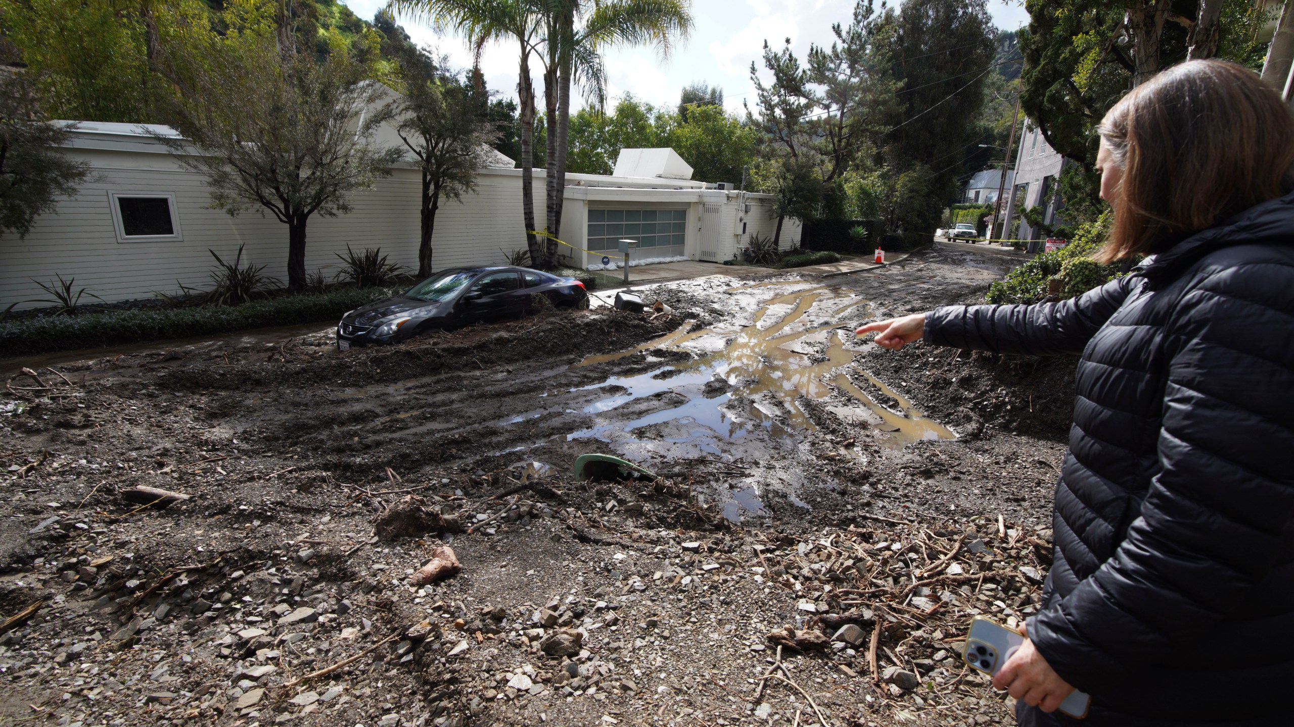 Local resident Jill Shinefield points to a mud filled street in her neighborhood in the Beverly Crest area of Los Angeles, on Wednesday, Feb. 7, 2024. A storm that parked itself over Southern California for days, unleashing historic downpours that caused hundreds of landslides, was moving out of the region after one final drenching Wednesday, but authorities warned of the continued threat of collapsing hillsides. (AP Photo/Richard Vogel)