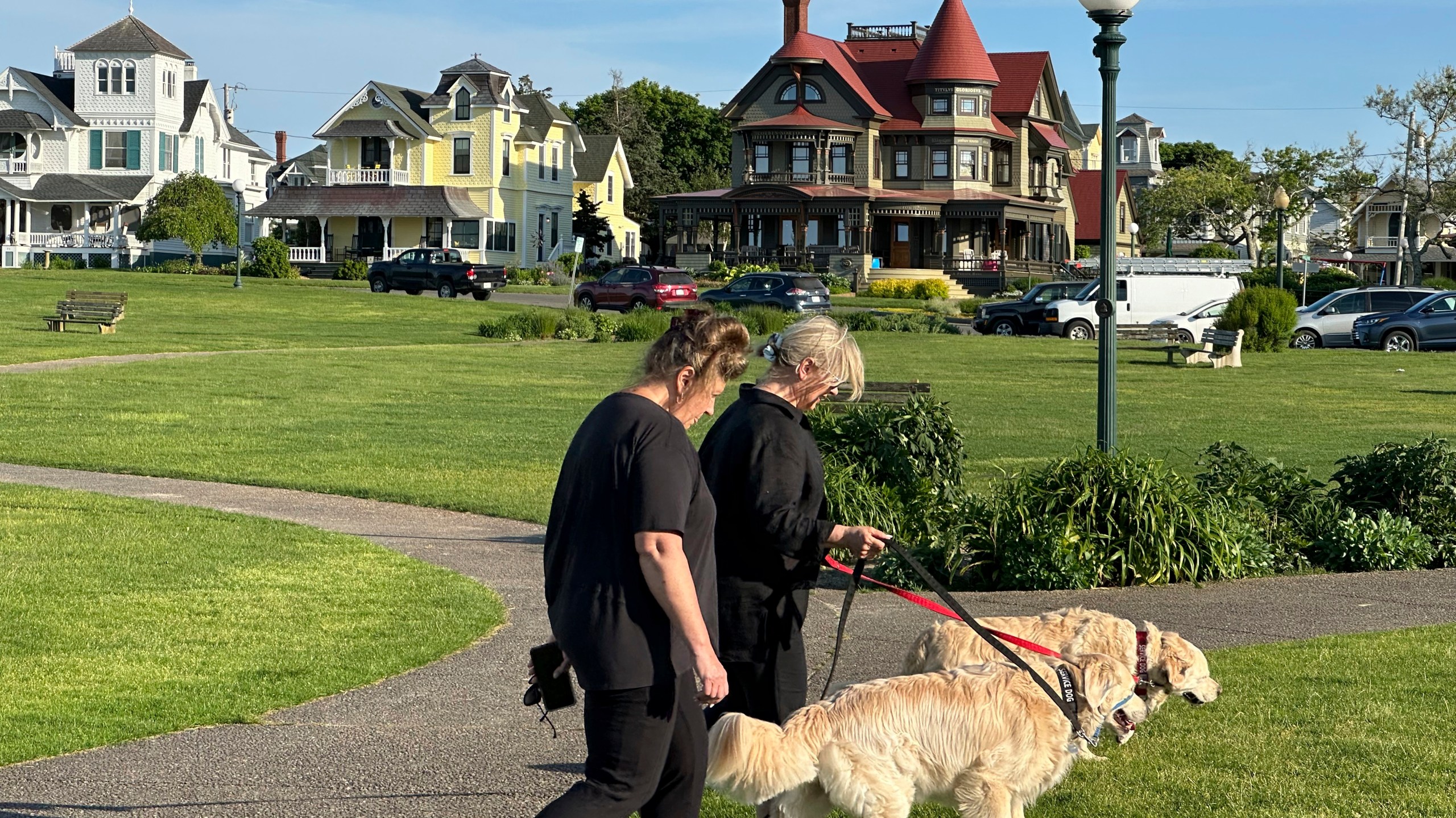 People walk dogs in front of homes, Tuesday, June 4, 2024, in Oak Bluffs, Massachusetts. High housing costs on Martha's Vineyard are forcing many regular workers to leave and are threatening public safety. (AP Photo/Nick Perry)