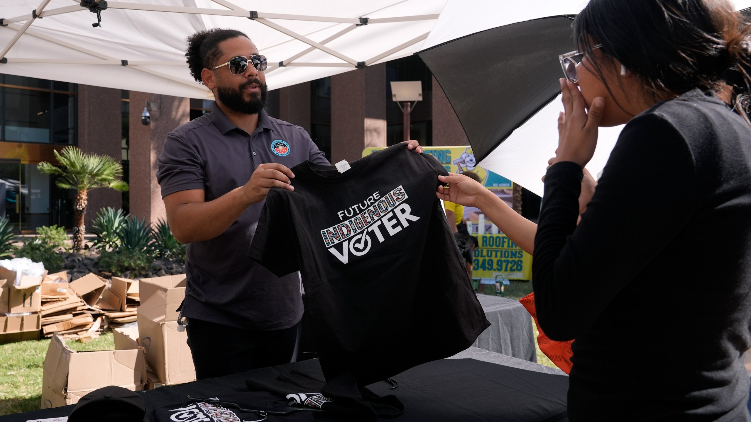 Alexander Castillo-Nunez, left, a civic engagement coordinator at the Inter Tribal Council of Arizona, Inc., shows a voter t-shirt at an Arizona Native Vote booth during an Indigenous Peoples' Day event Monday, Oct. 14, 2024, in Phoenix. (AP Photo/Ross D. Franklin)