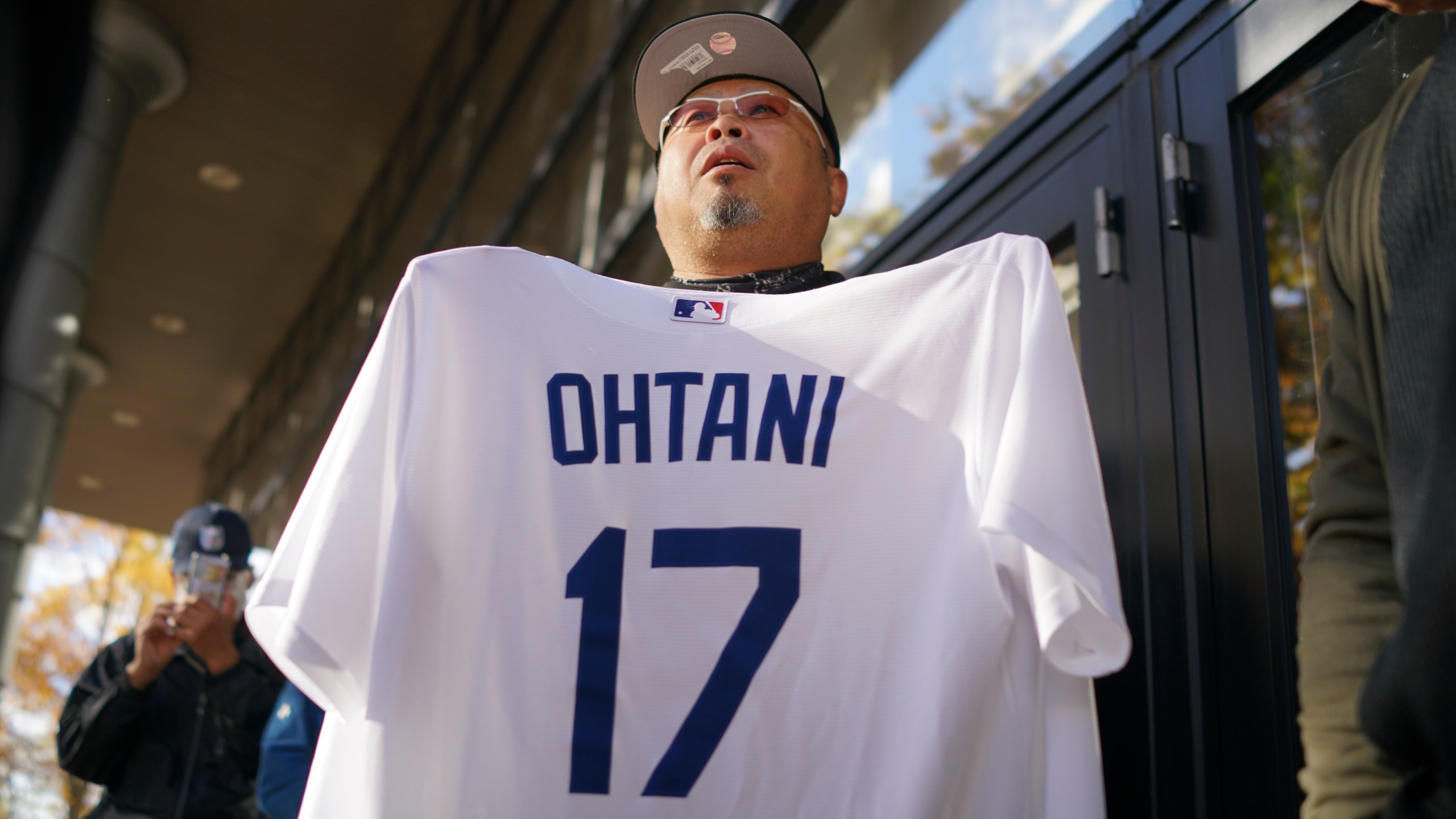 A man shows off a baseball jersey of Shohei Ohtani of the Los Angeles Dodgers as he and others wait in line to watch a live stream of Game 3 of the baseball World Series between the Dodgers and the New York Yankees, in a public viewing event in Oshu, northeastern Japan, the hometown of Ohtani, Tuesday, Oct. 29, 2024. (AP Photo/Eugene Hoshiko)