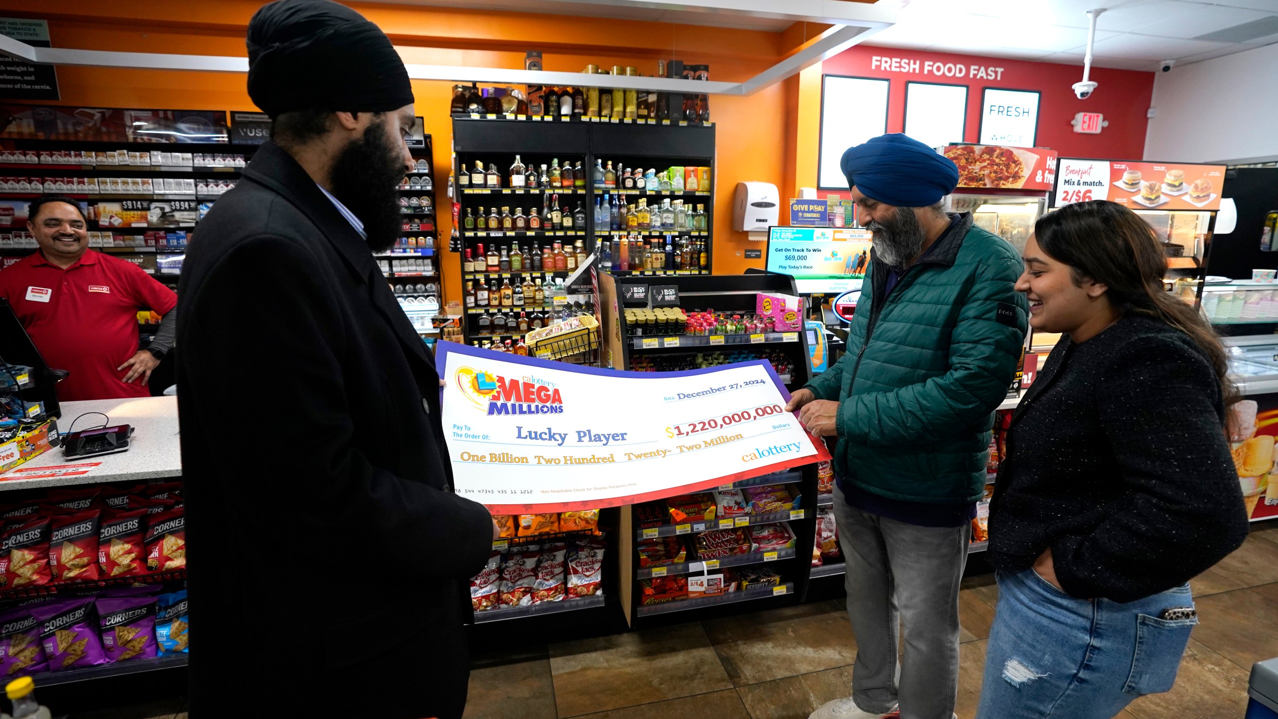 Ishar Gill, left, his father Jaspal Singh, center and his sister Jaspeet Gill right, look over the enlarged image of a check, Saturday, Dec. 28, 2024, that will be presented to the winner of $1.22 billion Mega Millions Lotto ticket that was purchased at their family's store in Cottonwood, Calif., Saturday, Dec. 28, 2024. (AP Photo/Rich Pedroncelli).