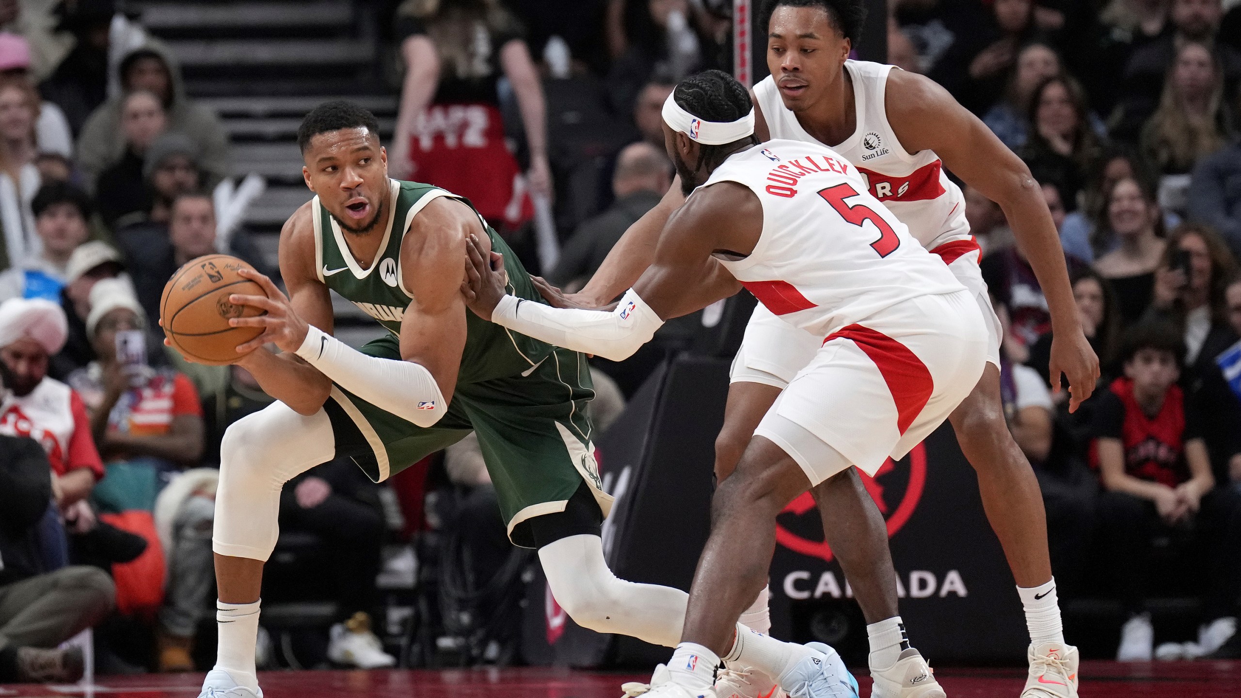 Milwaukee Bucks forward Giannis Antetokounmpo, left, looks to pass the ball while under pressure from Toronto Raptors guard Immanuel Quickley (5) and Scottie Barnes, right, during first-half NBA basketball game action in Toronto, Monday, Jan. 6, 2025. (Nathan Denette/The Canadian Press via AP)