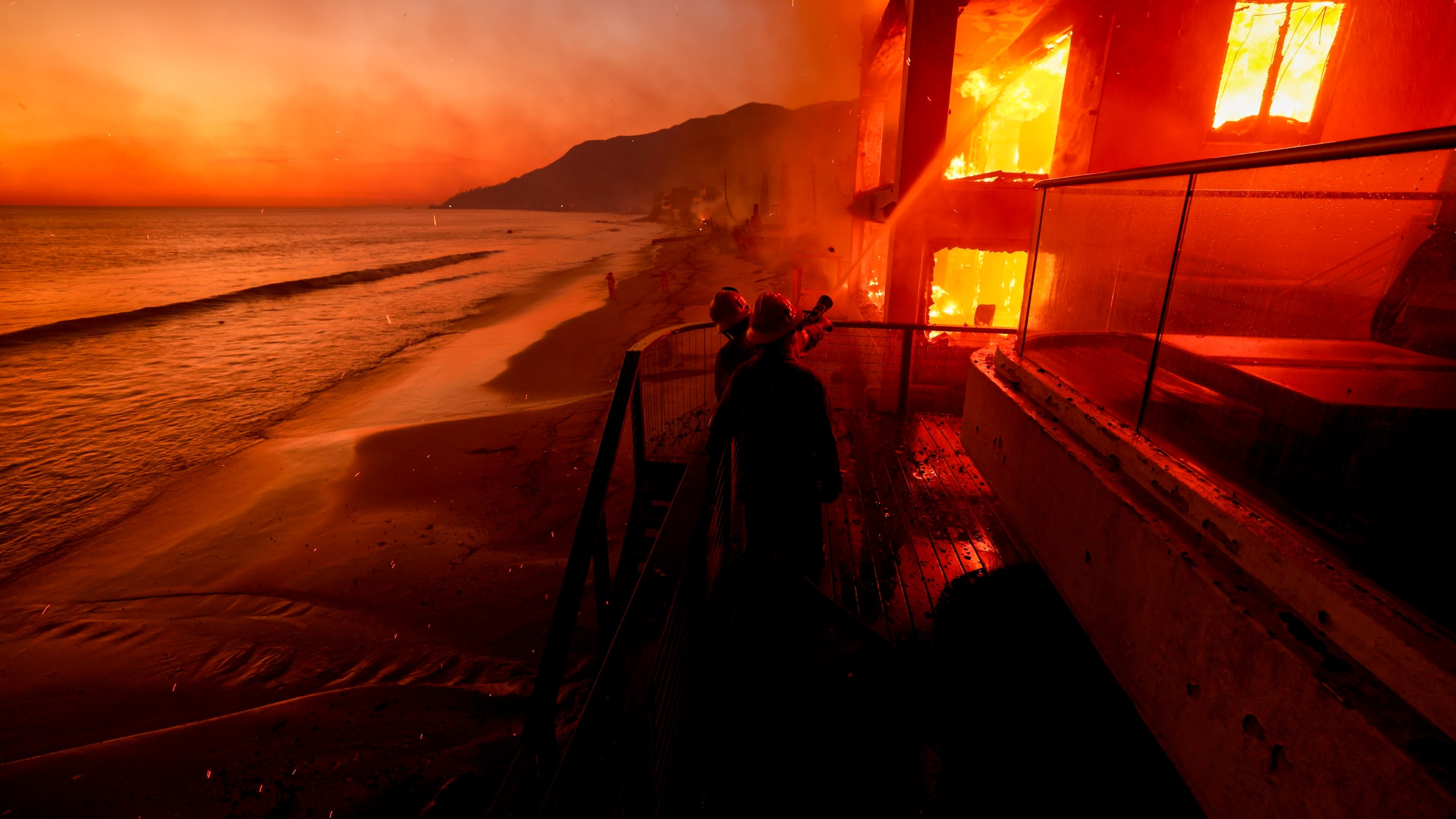 Firefighters work from a deck as the Palisades Fire burns a beachfront property Jan. 8, 2025, in Malibu, Calif. (AP Photo/Etienne Laurent)